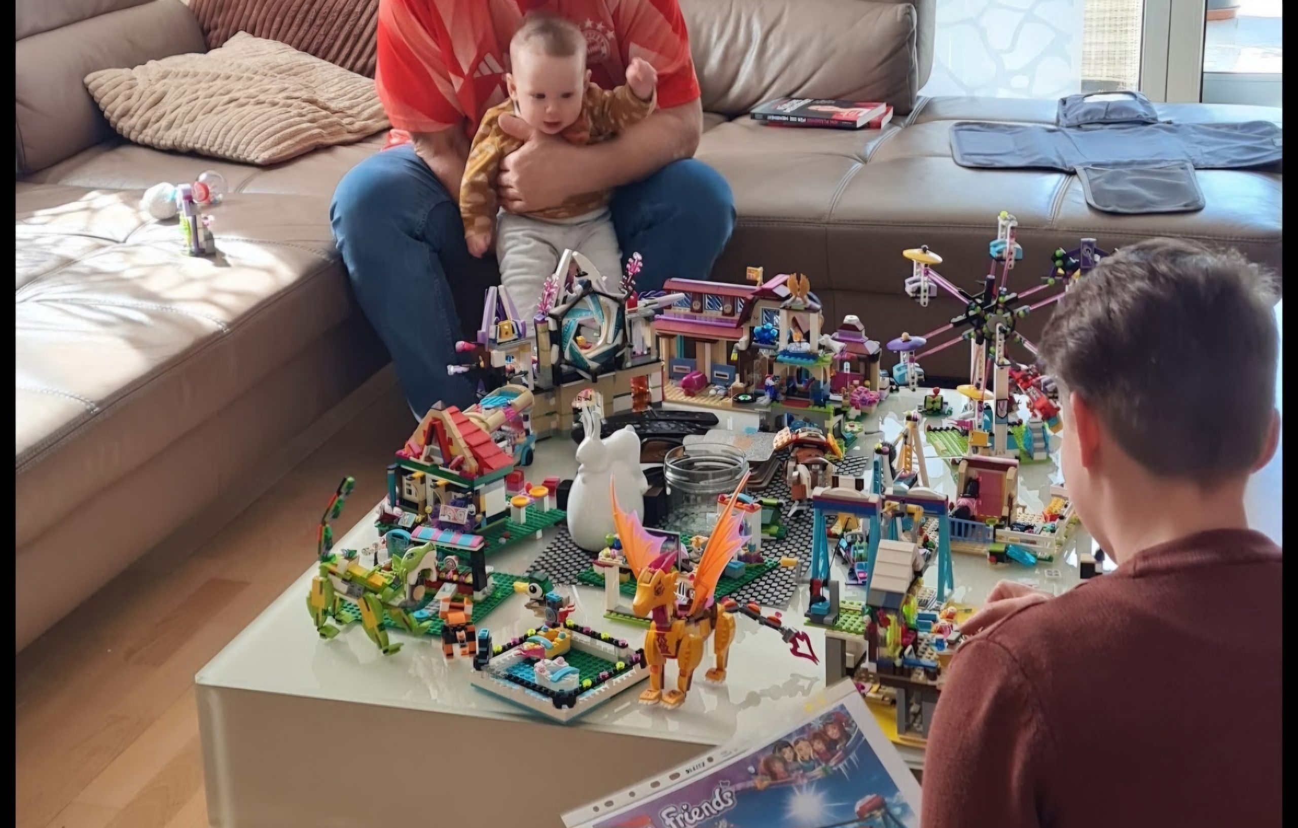 Ein Wohnzimmer. Der Wohnzimmertisch ist voll gestellt mit unterschiedlichen Legos. Im Hintergrund hält ein Mann das Baby Richtung Tisch. Es schaut voller Freude auf den Tisch.